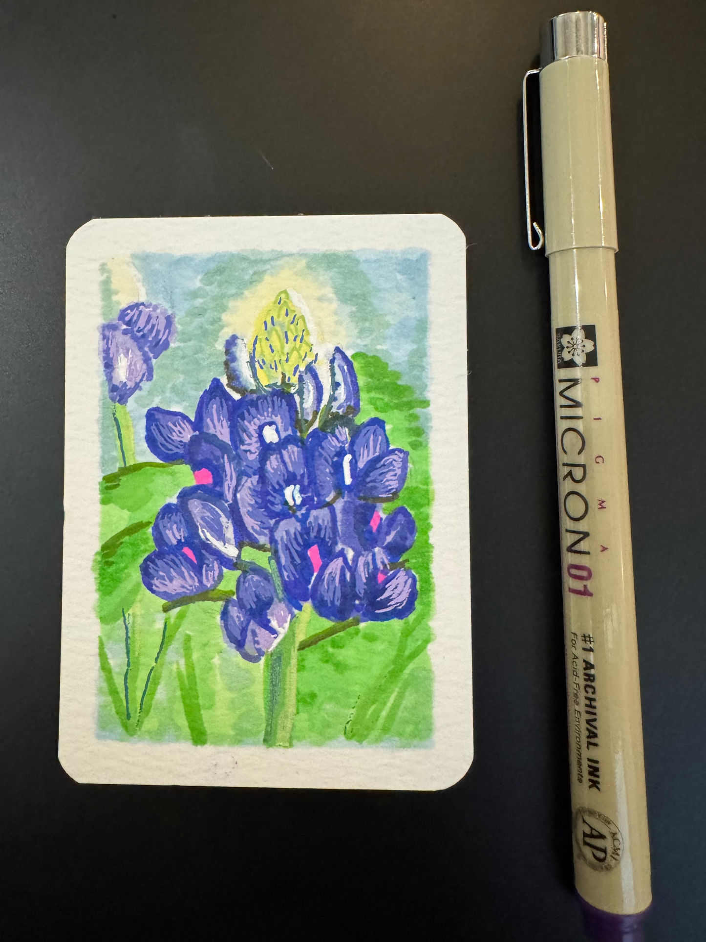 Small painted card with bluebonnet flowers next to a Micron pen on a dark surface