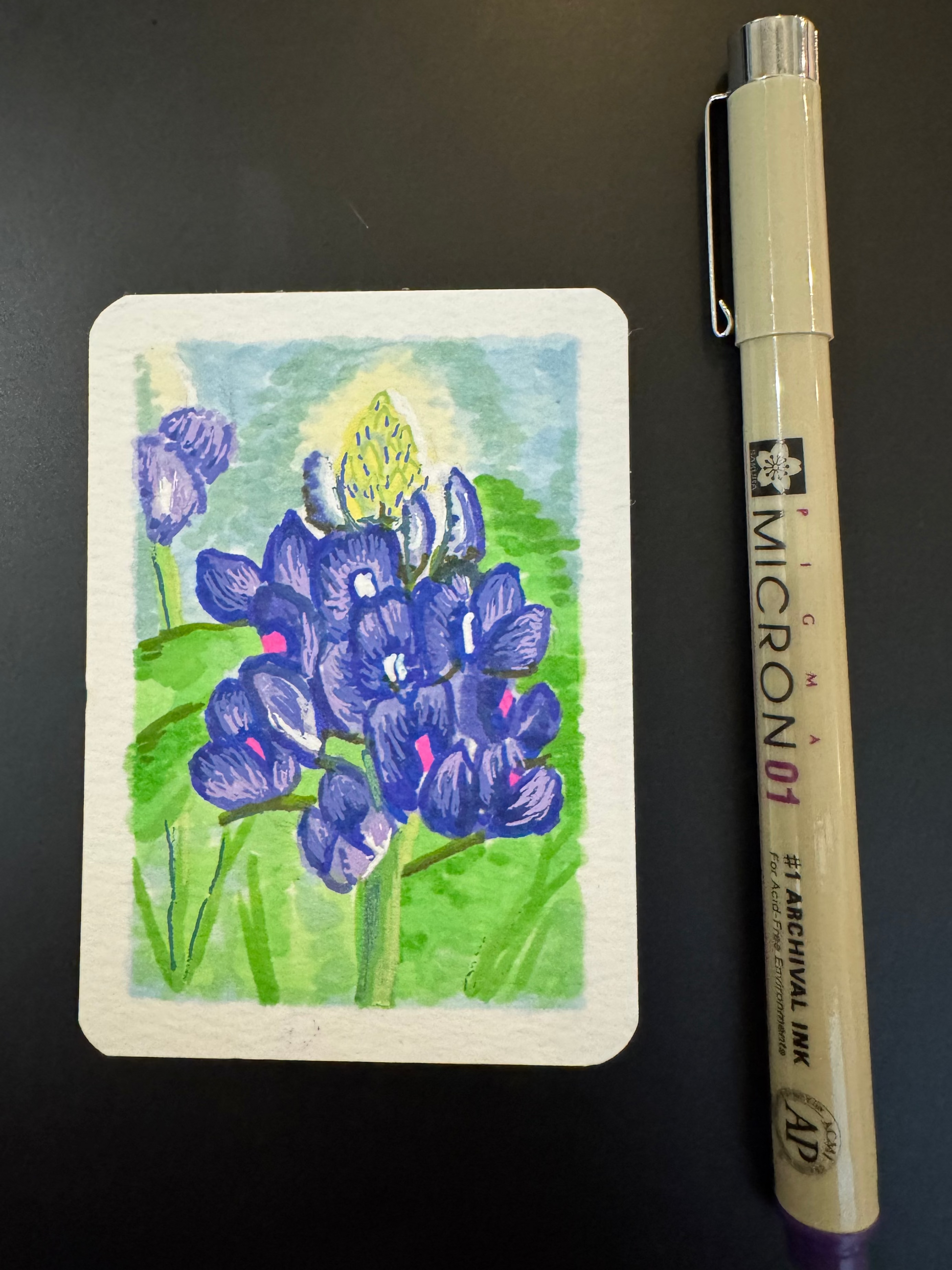 Small painted card with bluebonnet flowers next to a Micron pen on a dark surface